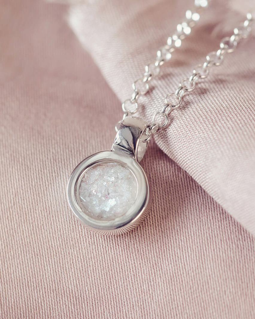 Sterling silver round necklace with a central section filled with memorial ashes and clear crushed crystals, displayed on a pink background