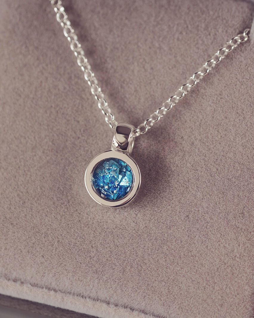 Sterling silver small round necklace with a central section filled with blue memorial ashes and crushed crystals, displayed on a beige fabric background.