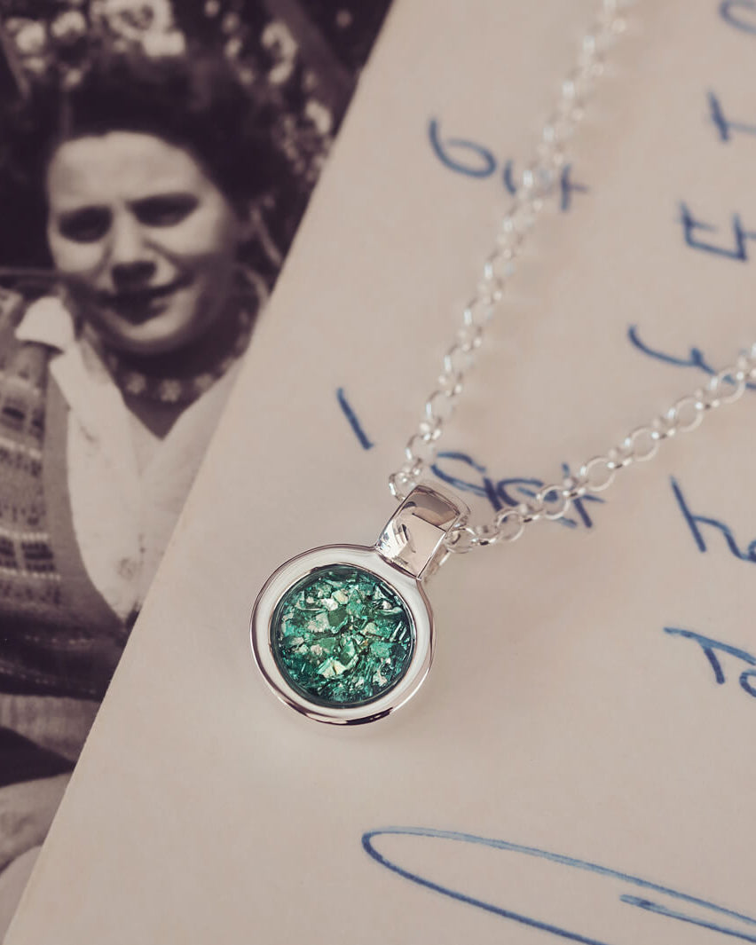 Sterling silver round necklace with a central section filled with memorial ashes and green crushed crystals, displayed on a background of a handwritten message and photograph