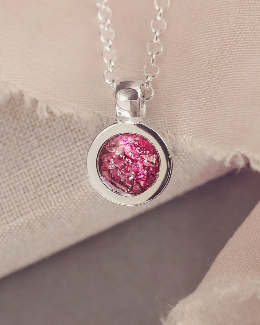 Sterling silver round necklace with a central section filled with memorial ashes and pink crushed crystals, displayed on a beige background