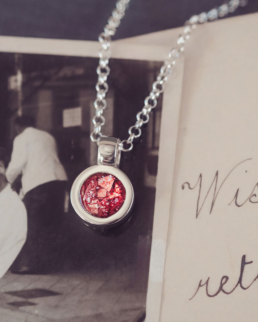 Sterling silver round necklace with a central section filled with memorial ashes and red crushed crystals, displayed on a background of a handwritten message and photograph