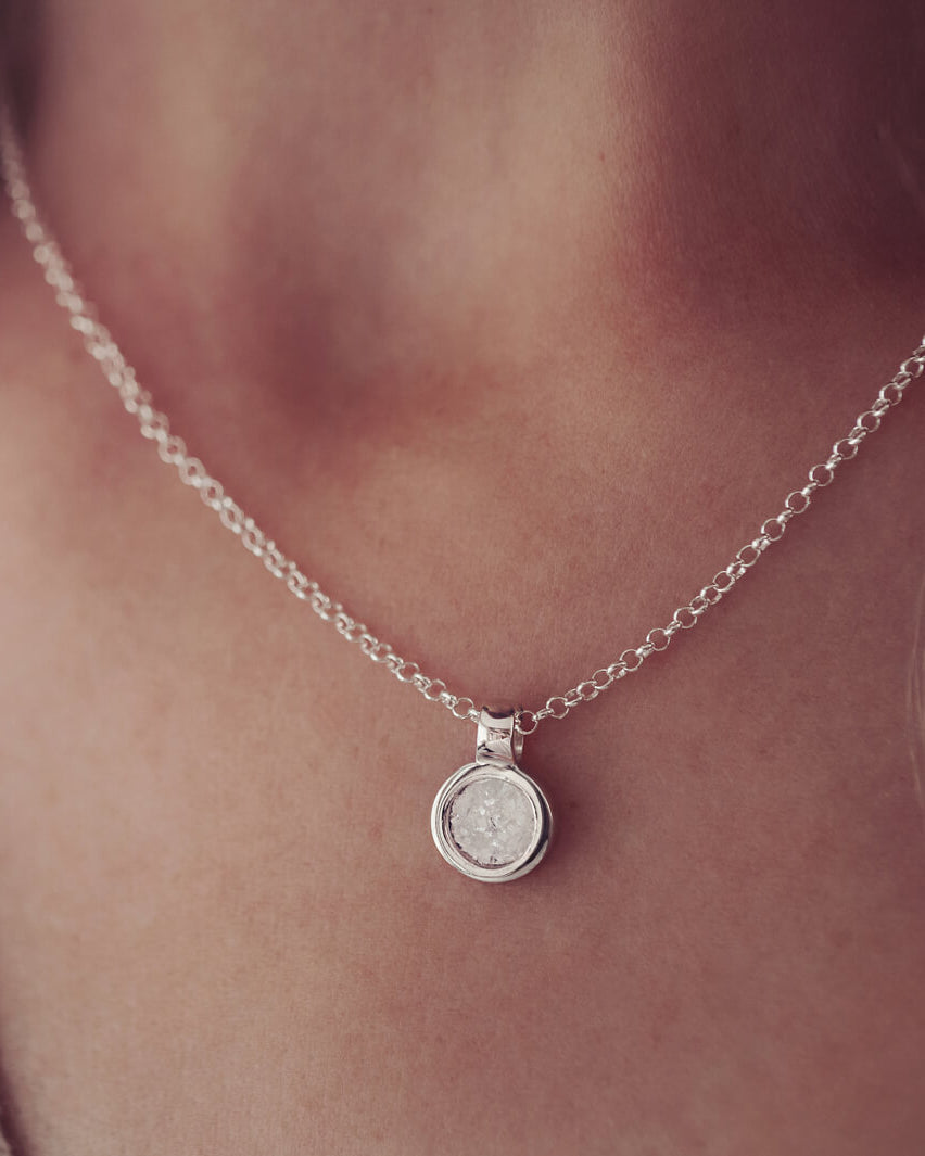 Lady wearing a small round cremation ashes necklace in the colour clear. Blonde hair and beige cardigan just visible in the image.