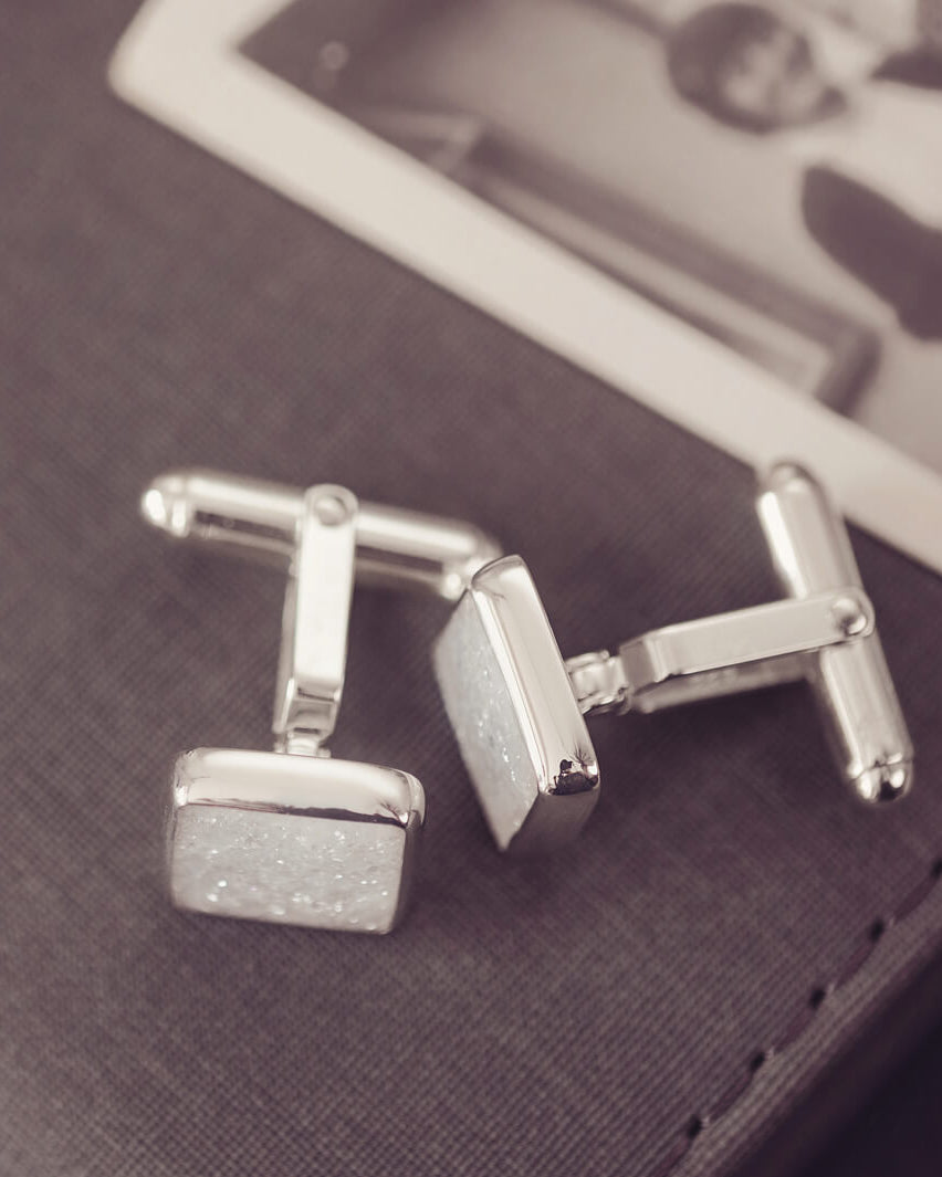 Square silver cufflinks containing cremation ashes, clear crystal and resin laying on their side on a black box