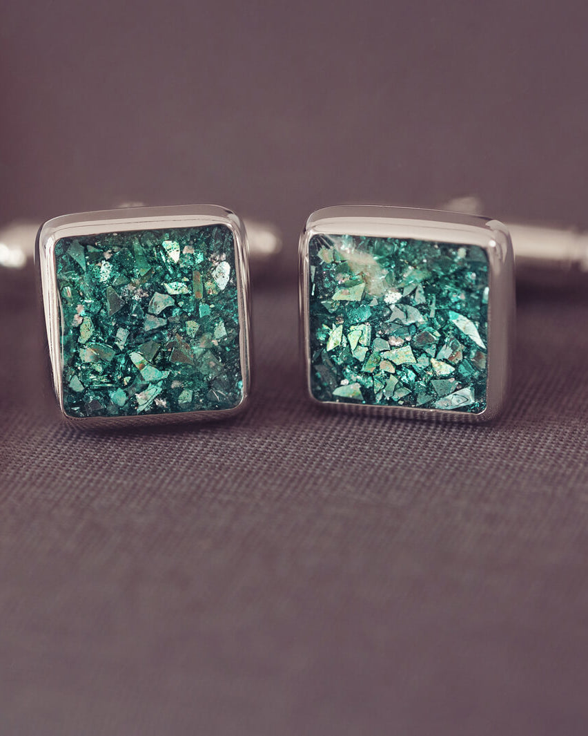 Square silver cufflinks containing cremation ashes, green crystal and resin sat on a dark grey background