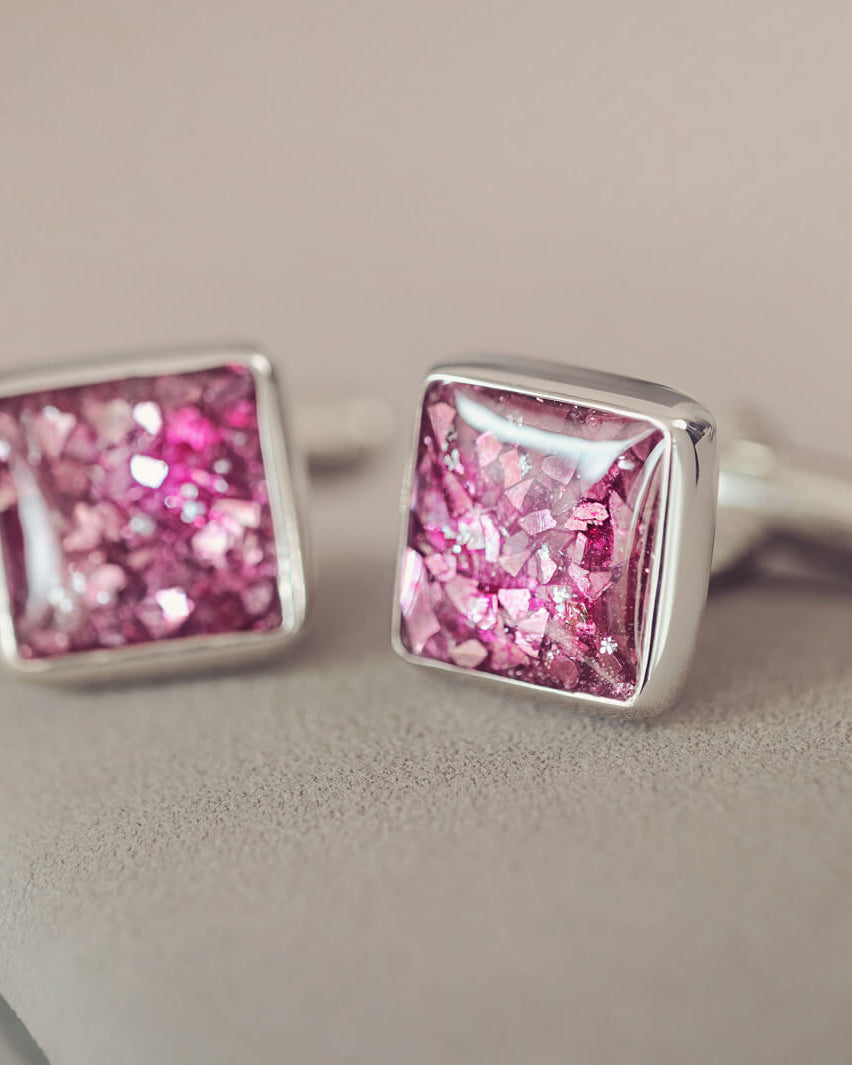Square silver cufflinks containing cremation ashes, pink crystal and resin sat on a light grey background