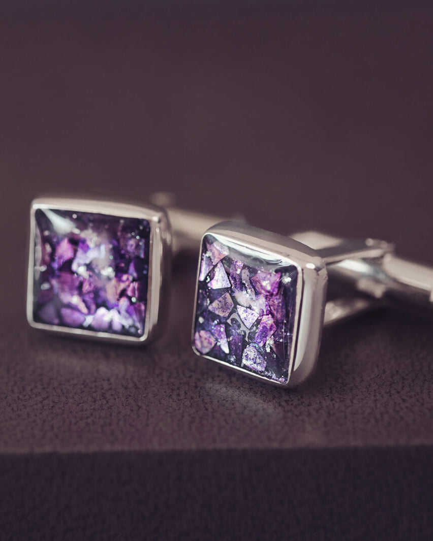 Square silver cufflinks containing cremation ashes, purple crystal and resin sat on a dark grey background