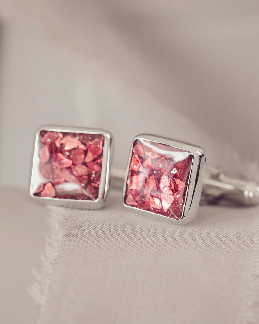Square silver cufflinks containing cremation ashes, red crystal and resin sat on a beige fabric background