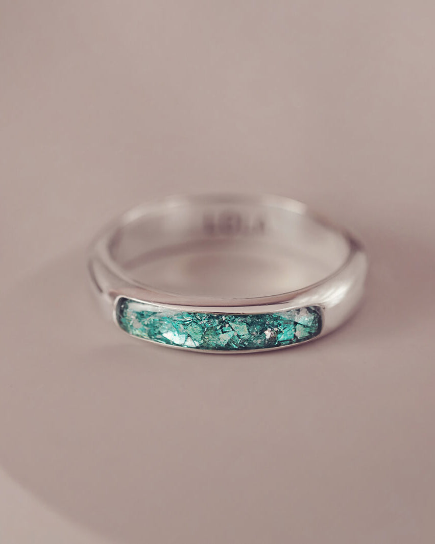 Sterling silver womens ring band with a central section filled with  memorial ashes and green crushed crystals, displayed on a pale pink background
