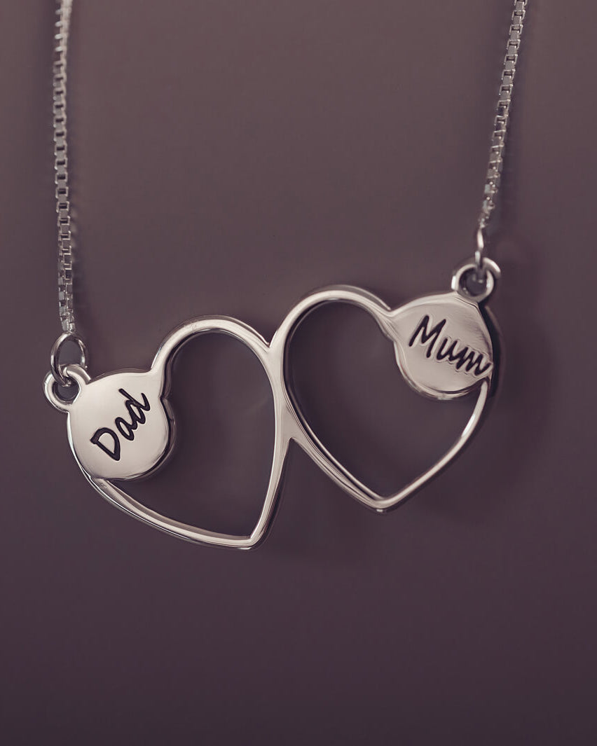 A delicate silver necklace featuring two intertwined heart-shaped pendants, engraved with a three character name on each heart. Displayed against a neutral background.