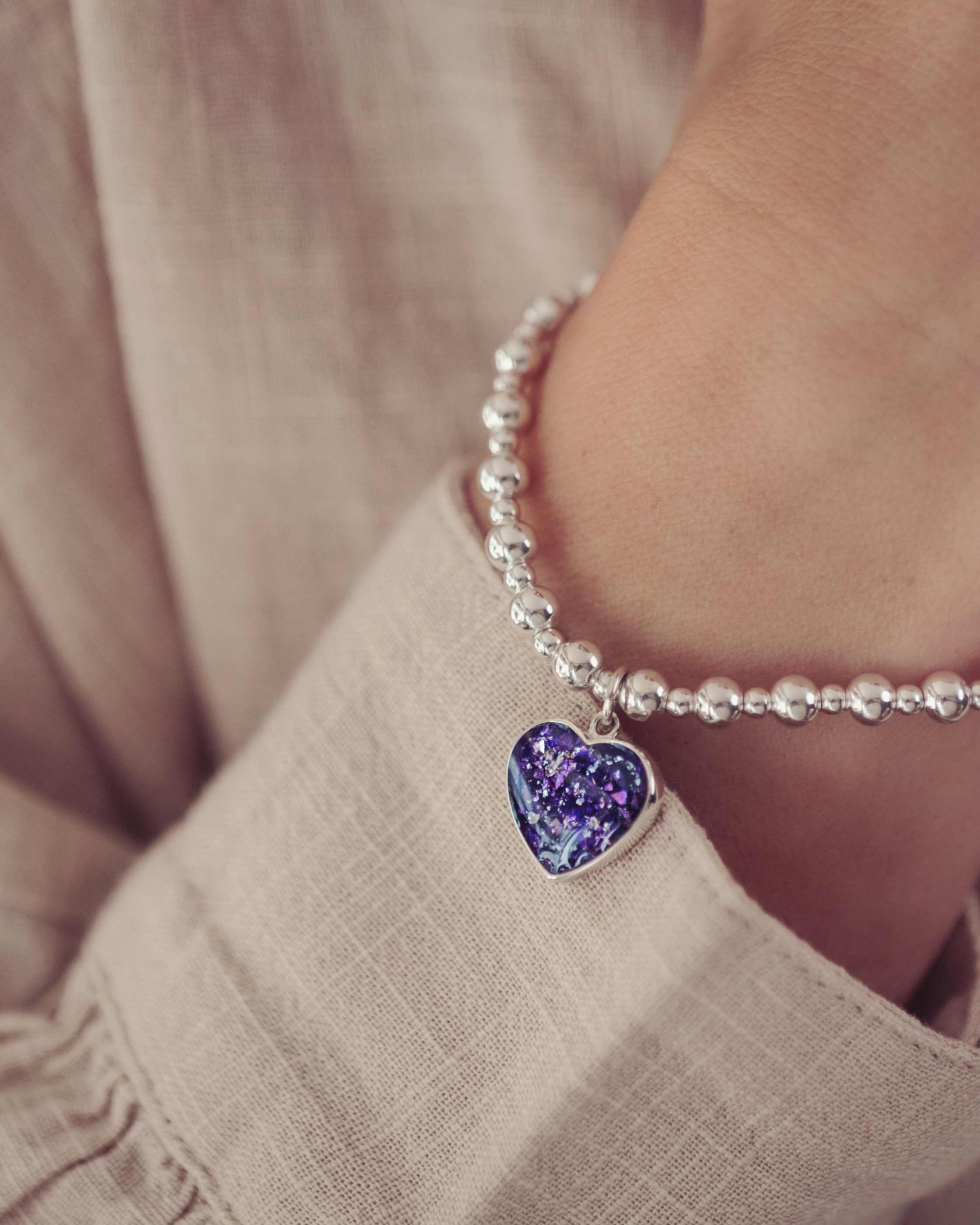 The image shows a close-up of a person's wrist adorned with a bracelet made of small silver beads. A small, heart-shaped charm hangs from the bracelet, filled with a deep purple, glittery resin and cremation ashes. The person is wearing a light, textured fabric, and the soft lighting emphasises the sentimental and elegant nature of the bracelet.