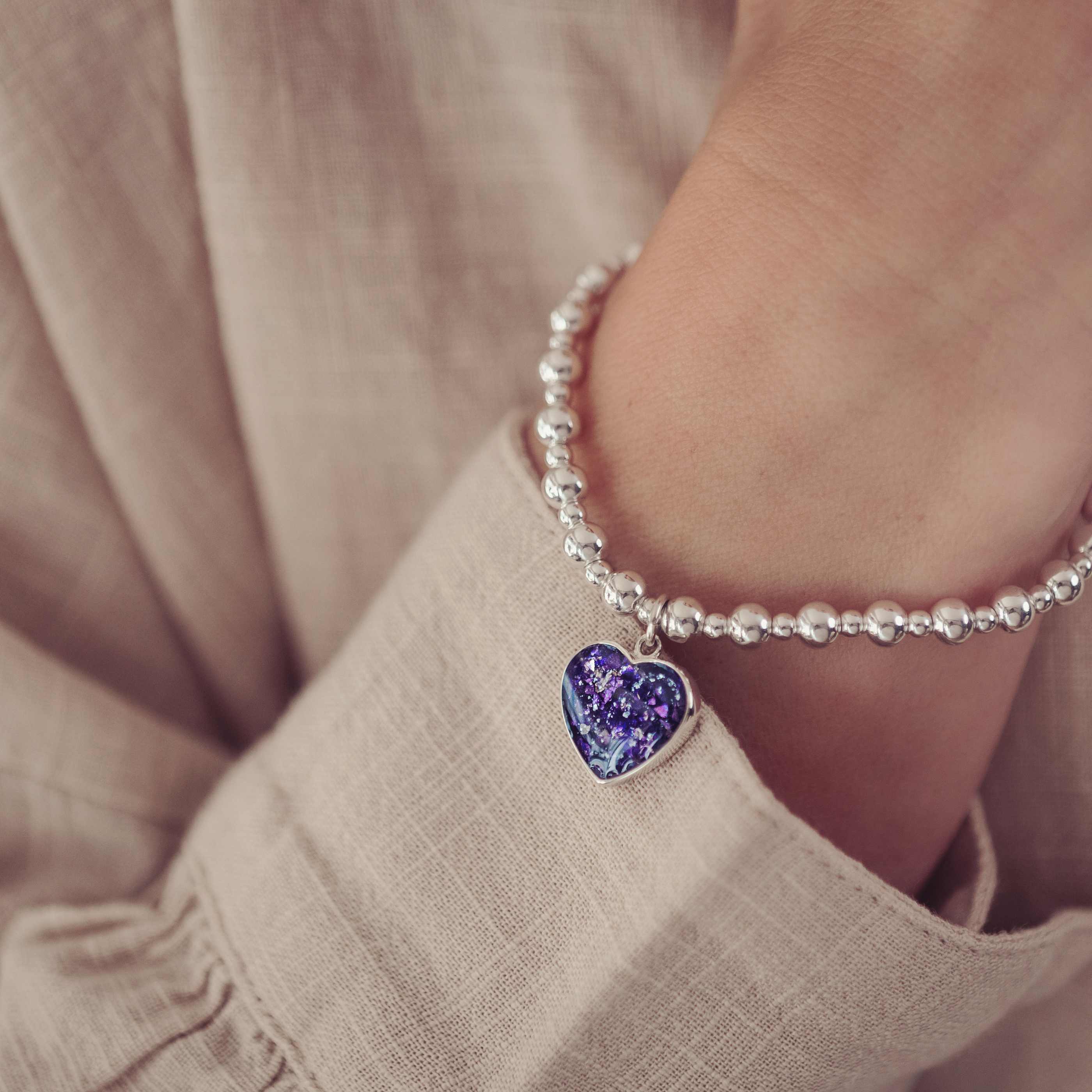The image shows a close-up of a person's wrist adorned with a bracelet made of small silver beads. A small, heart-shaped charm hangs from the bracelet, filled with a deep purple, glittery resin and cremation ashes. The person is wearing a light, textured fabric, and the soft lighting emphasises the sentimental and elegant nature of the bracelet.