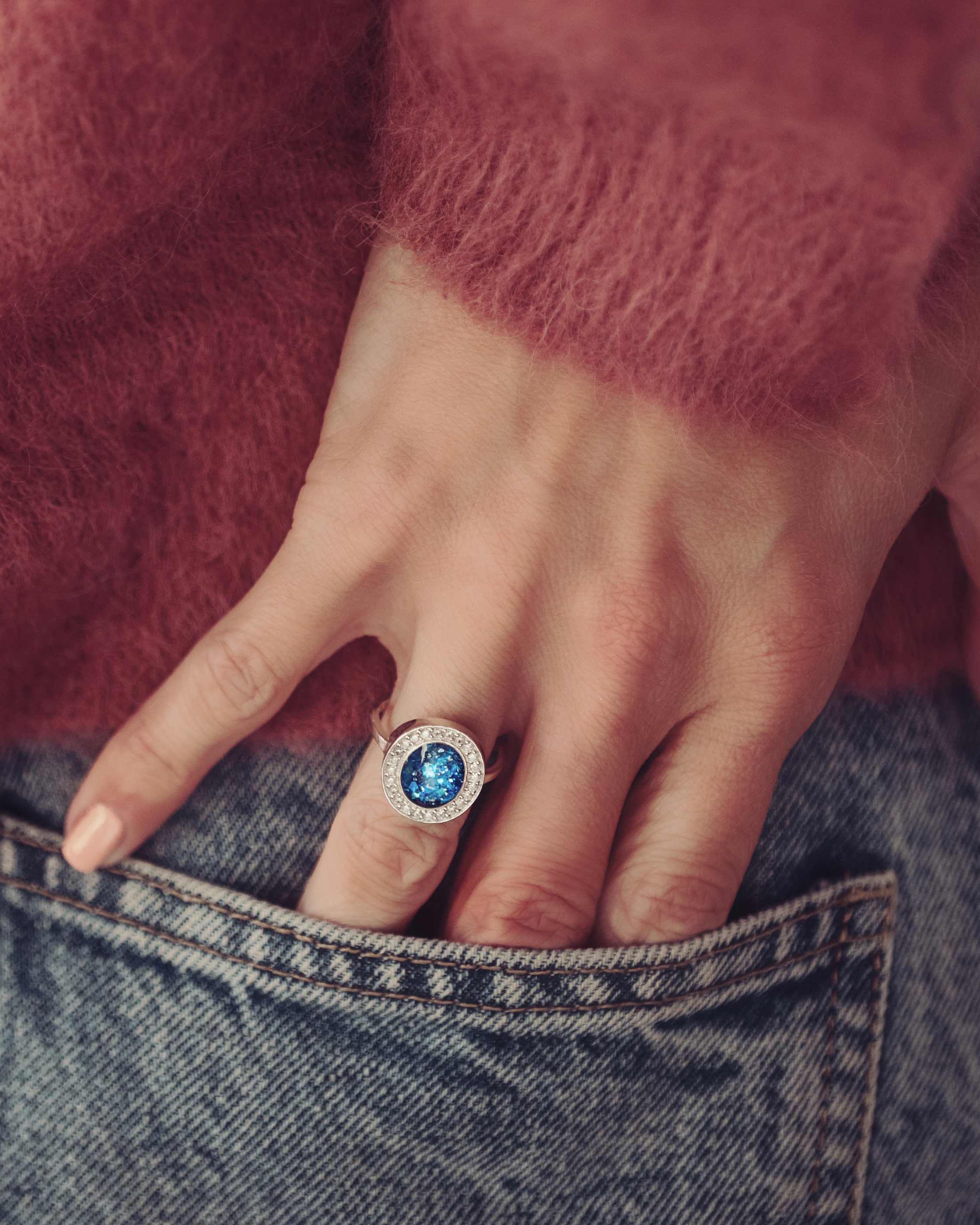 The image shows a close-up of a person’s hand resting near their back pocket, with their fingers partially tucked into blue denim jeans. They are wearing a large, silver ring featuring a vibrant blue stone containing cremation ashes and resin, surrounded by a halo of smaller stones. The person is dressed in a soft, textured, rose-coloured sweater, and the rich colours of the sweater and the blue stone create a striking contrast. The ring stands out as a bold and elegant piece.