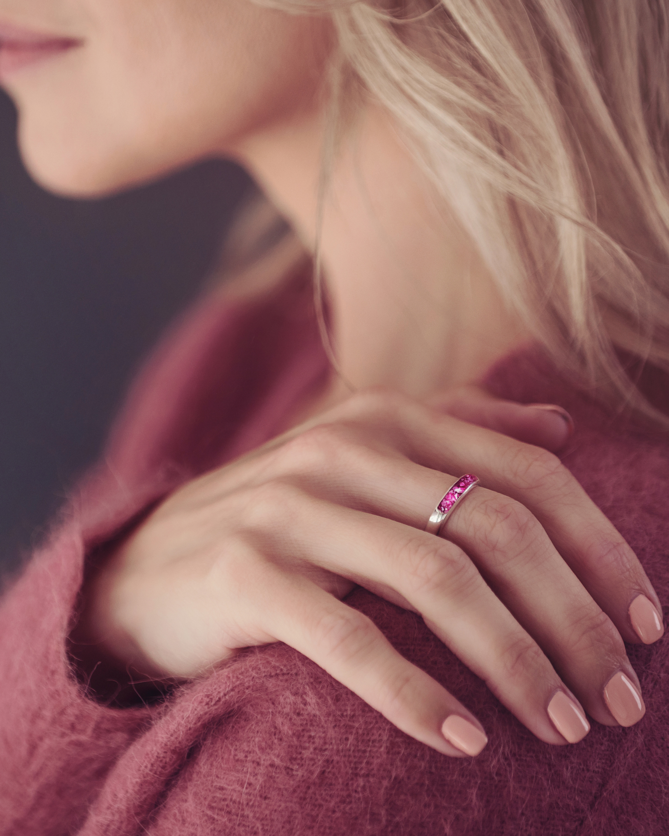 The image shows a close-up of a person’s hand resting on their shoulder. They are wearing a silver ring with a band of bright pink stone, containing cremation ashes and resin. The person is dressed in a soft, textured, rose-coloured sweater that complements the colour of the stone. Their nails are neatly manicured with a neutral polish. The soft lighting highlights the ring, drawing attention to its vibrant colour and elegant design, suggesting that it holds sentimental or memorial significance.
