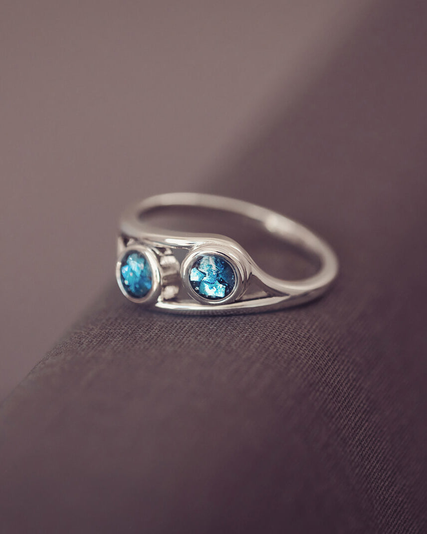 sterling silver ring with two circles created with blue coloured crystal and cremation ashes laying on a dark grey background
