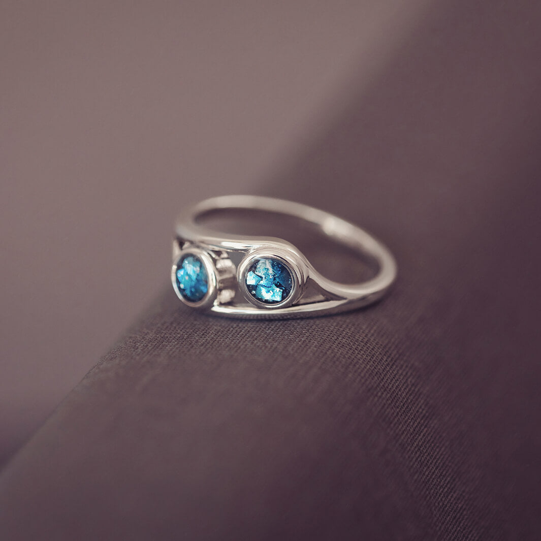 sterling silver ring with two circles created with blue coloured crystal and cremation ashes laying on a dark grey background