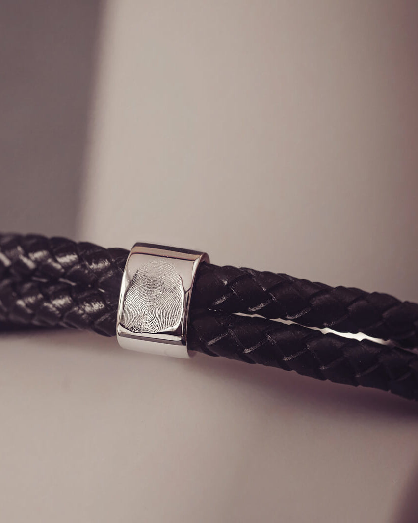 Black braided leather bracelet featuring a silver charm engraved with a fingerprint, displayed in a grey jewellery box
