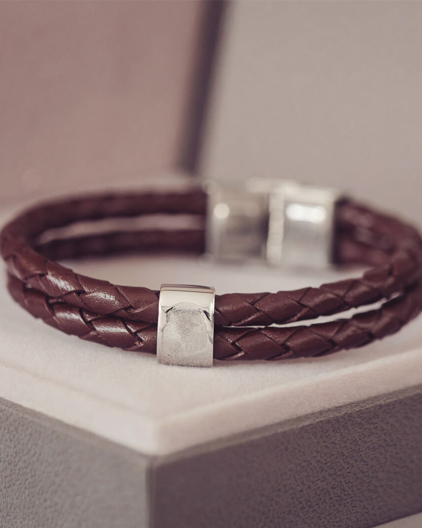 Brown braided leather bracelet featuring a silver charm engraved with a fingerprint, displayed in a grey jewellery box