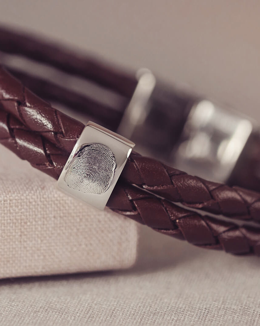 Brown braided leather bracelet featuring a silver charm engraved with a fingerprint, displayed in a grey jewellery box