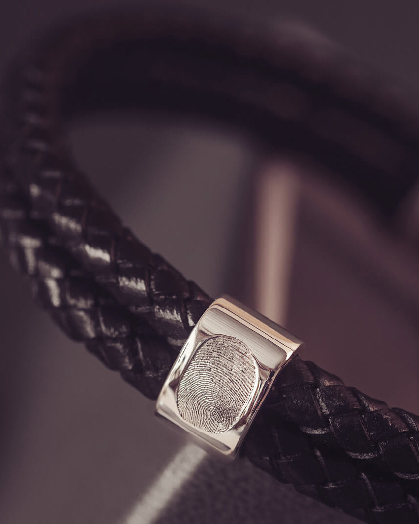 Black braided leather bracelet featuring a silver charm engraved with a fingerprint, displayed in a grey jewellery box