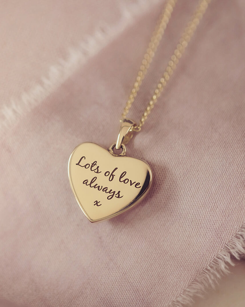Reverse of the small gold heart necklace with engraving in our standard font laying on a beige fabric background
