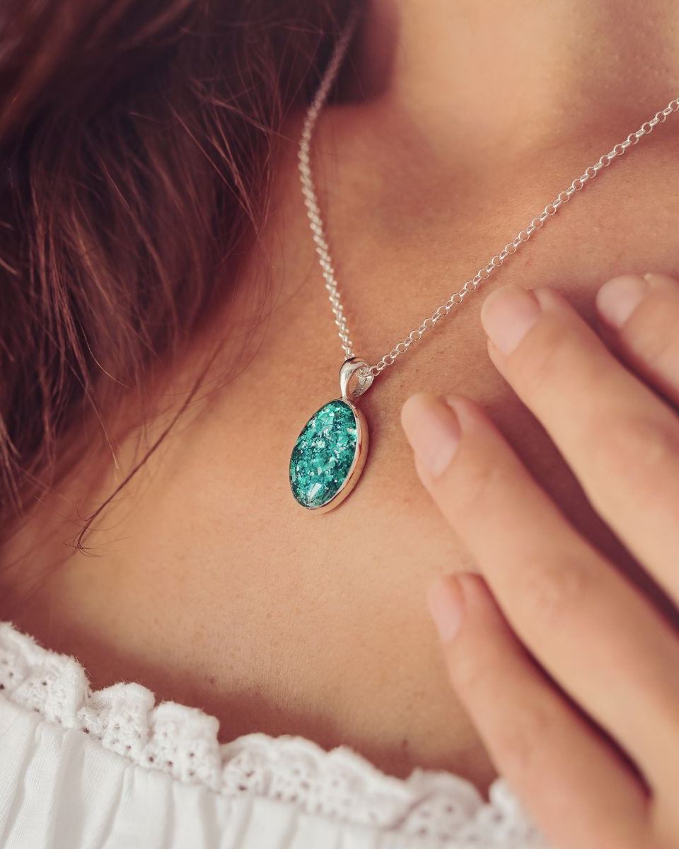 Neckline of a lady with brown hair wearing a Cremation Ashes Oval Necklace, filled with ashes and green crushed crystal. The lady is wearing a white top and is holding her hand towards the necklace