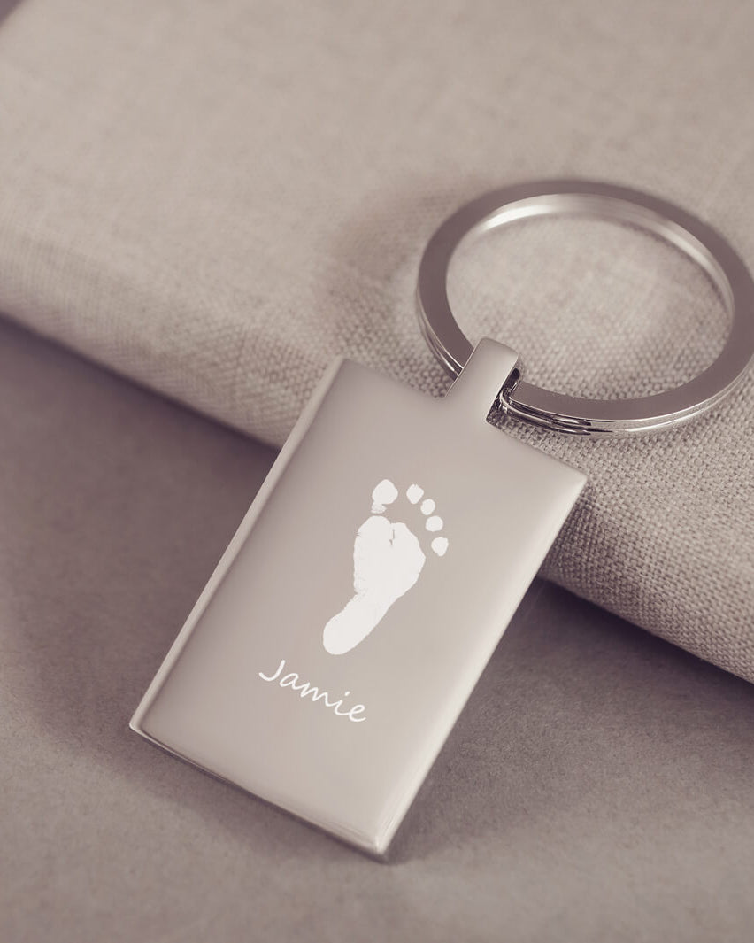 A stainless steel rectangular keyring, engraved with one footprint and name, leaning on a fabric background