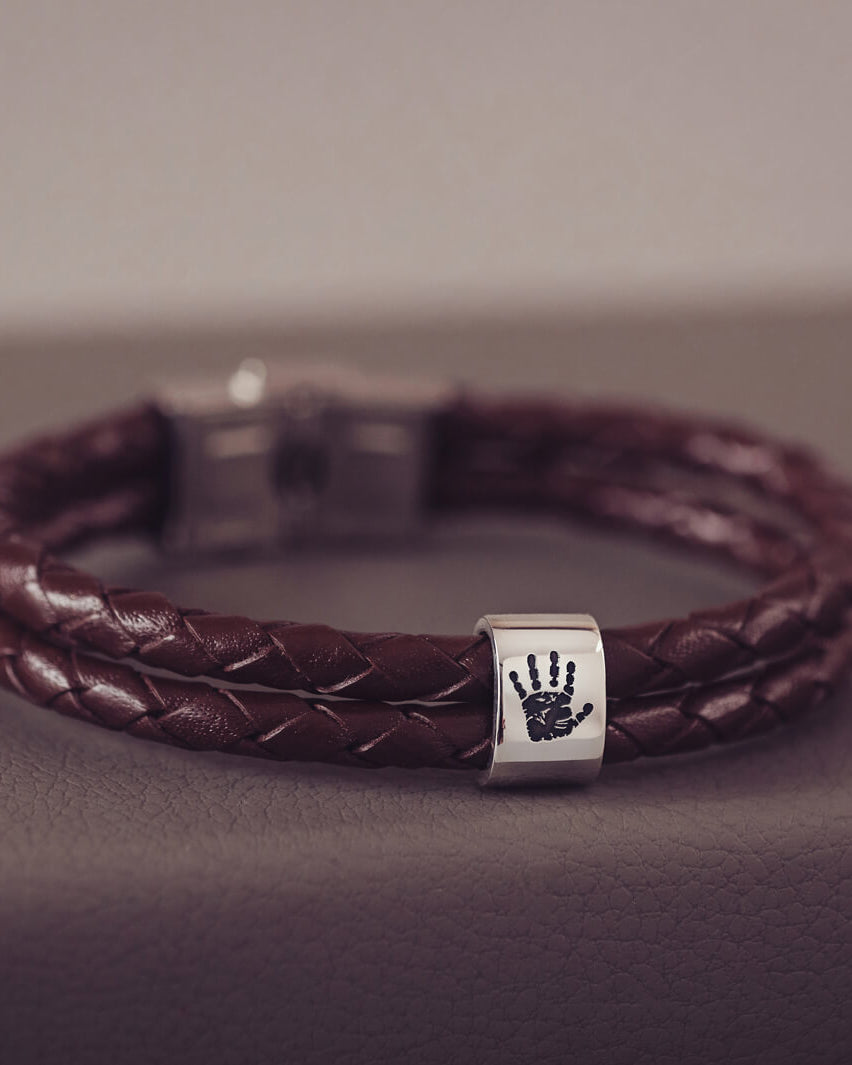 Brown braided leather bracelet featuring a sterling silver charm engraved with a black handprint, displayed on a soft grey background.