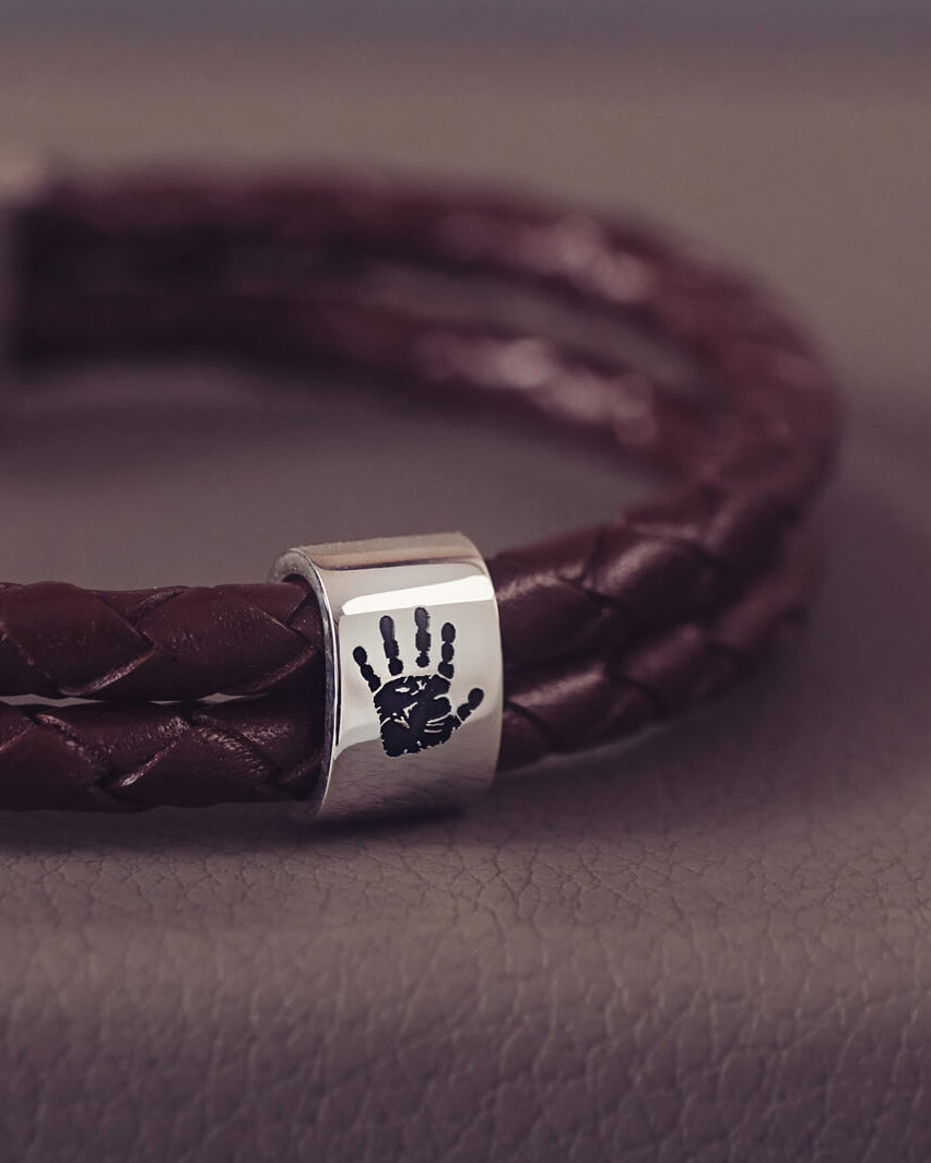 Brown braided leather bracelet featuring a sterling silver charm engraved with a black handprint, displayed on a soft grey background.