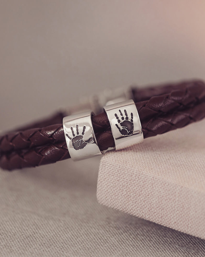 Brown braided leather bracelet featuring two sterling silver charms, each engraved with a black handprint, displayed on a soft grey background.