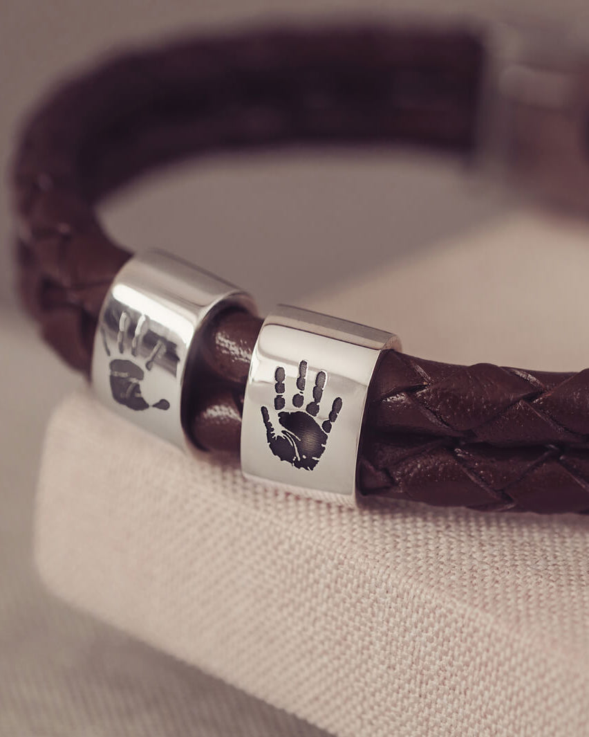 Brown braided leather bracelet featuring two sterling silver charms, each engraved with a black handprint, displayed on a soft grey background.