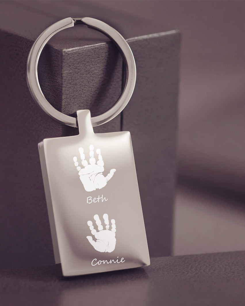 A stainless steel rectangular keyring resting on a dark grey jewellery box, with two handprints and names engraved onto the keyring