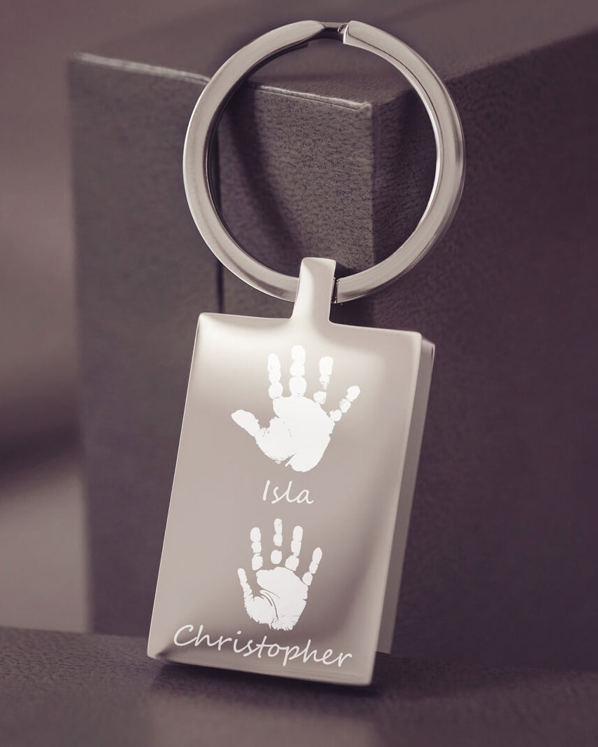 A stainless steel rectangular keyring resting on a dark grey jewellery box, with two handprints and names engraved onto the keyring