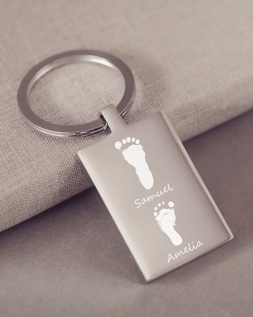A stainless steel rectangular keyring, engraved with two footprints and names, leaning on a fabric background