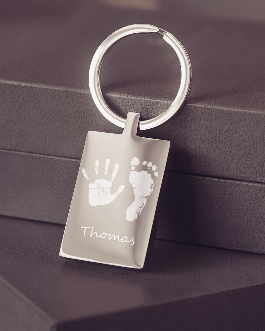 A stainless steel keyring, engraved with a handprint, footprint and name, leaning against a grey jewellery box