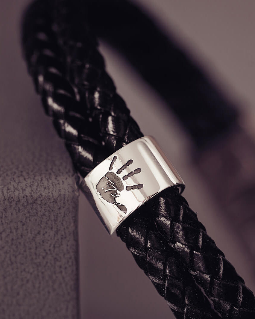 Black braided leather bracelet featuring a sterling silver charm engraved with a black handprint, displayed on a soft grey background.