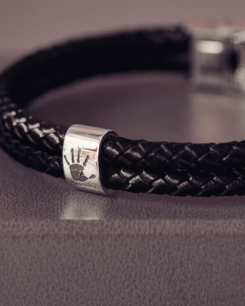Black braided leather bracelet featuring a sterling silver charm engraved with a black handprint, displayed on a soft grey background.