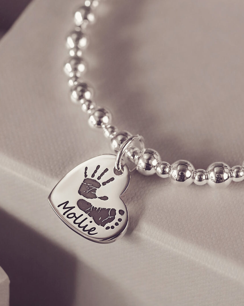 Silver beaded bracelet featuring a heart-shaped charm engraved with a handprint and footprint, along with the name 'Mollie,' displayed on a grey surface