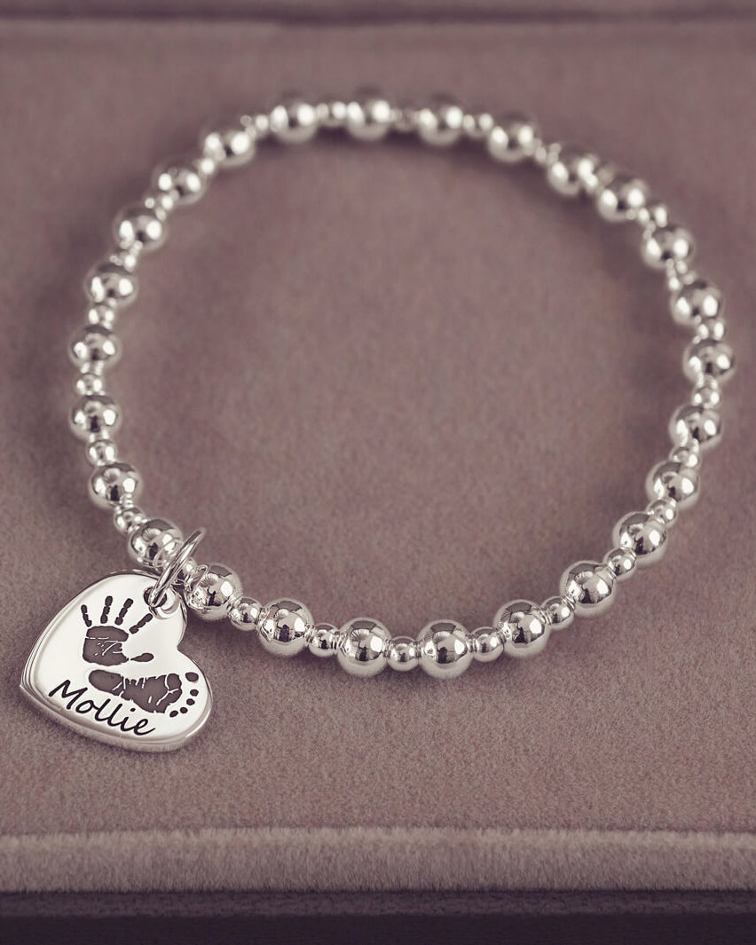 Silver beaded bracelet featuring a heart-shaped charm engraved with a handprint and footprint, along with the name 'Mollie,' displayed on a soft beige surface