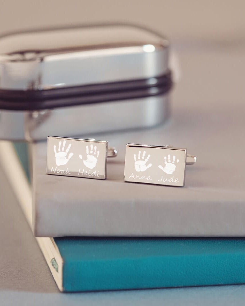 stainless steel rectangular cufflinks with two handprints and names engraved onto each cufflink. Displayed on a grey and blue background, with a chrome cufflink case in the background