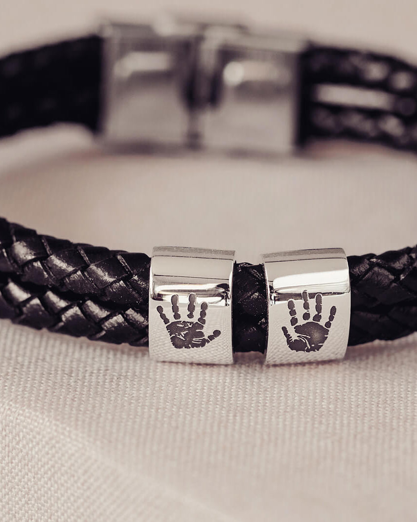 Black braided leather bracelet featuring two sterling silver charms, each engraved with a black handprint, displayed on a soft grey background.