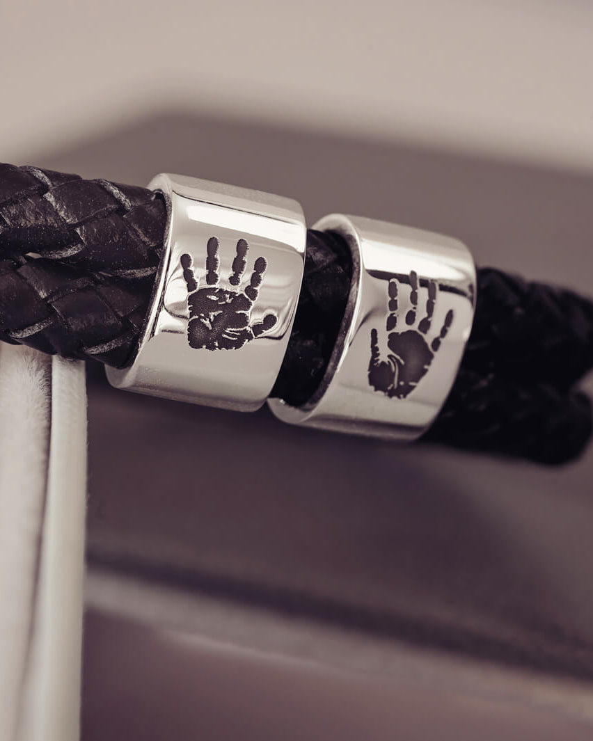 Black braided leather bracelet featuring two sterling silver charms, each engraved with a black handprint, displayed on a soft grey background.