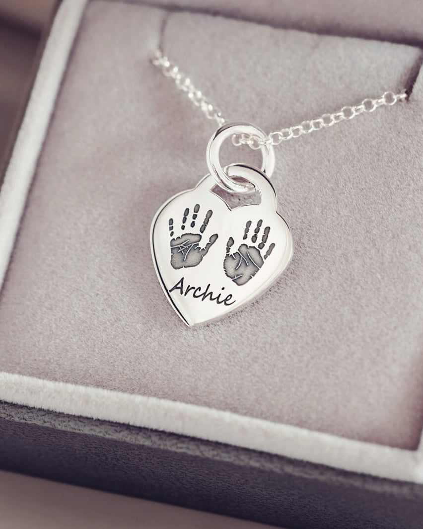 A silver lock heart necklace, on a silver chain, with two black handprint and the name 'Archie' impressed into the front of the necklace. Displayed in a grey jewellery box