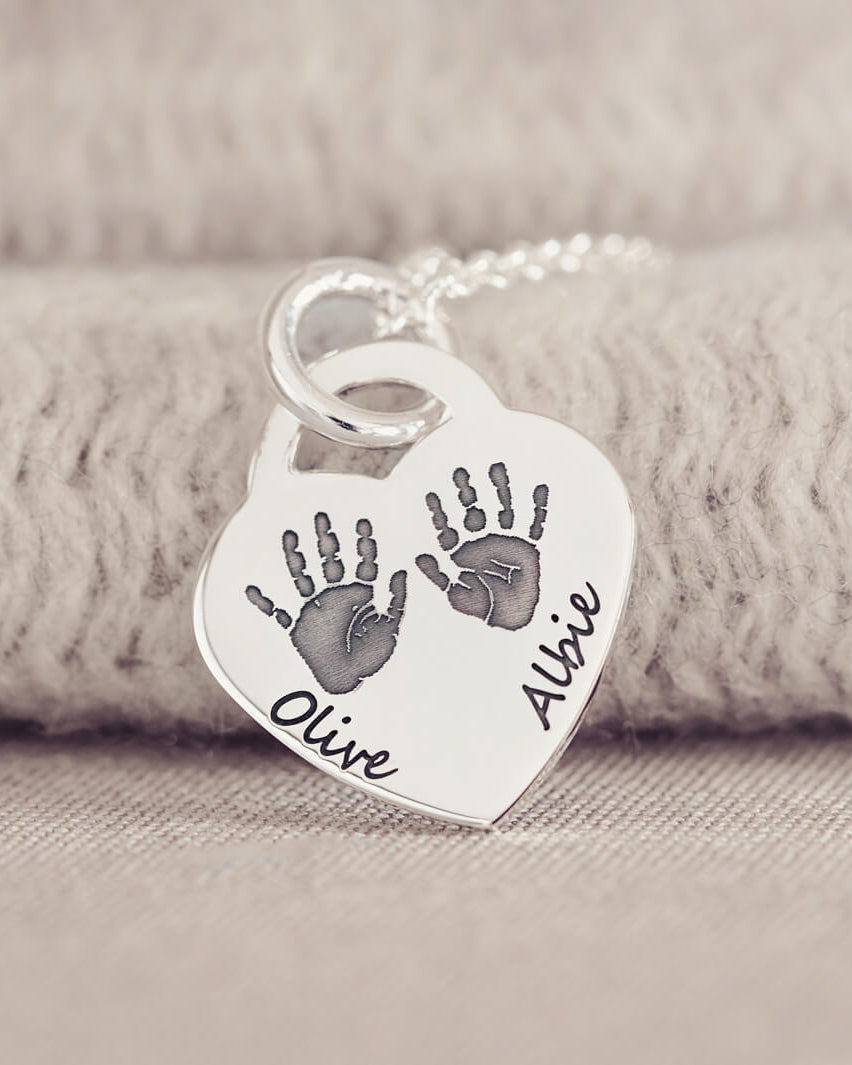 A silver heart shaped necklace, on a silver chain, with two handprints and names Olive and Albie engraved onto the front of the pendant. Displayed on a beige fabric background