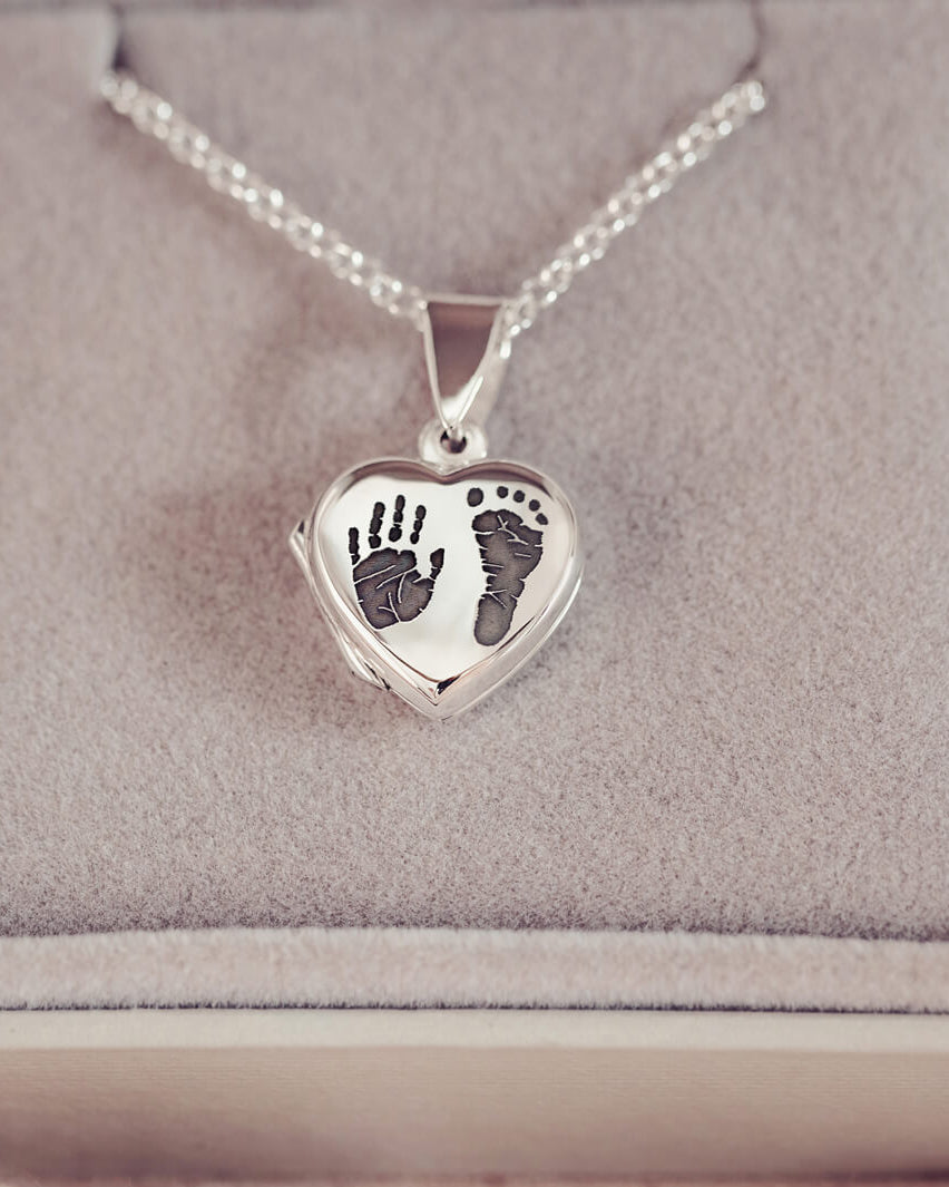 A silver heart shaped hinged locket, on a silver belcher chain, with a dark grey hand and footprint engraved into the front of the locket pendant. Displayed in a grey jewellery box