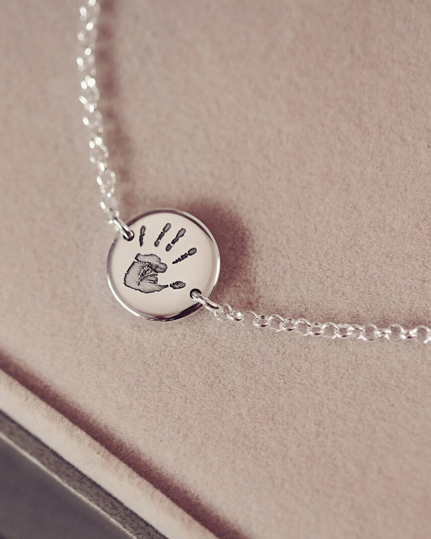 A round silver pendant bracelet featuring a detailed engraved handprint, displayed on a delicate silver chain. The necklace rests in an elegant, soft velvet jewellery box