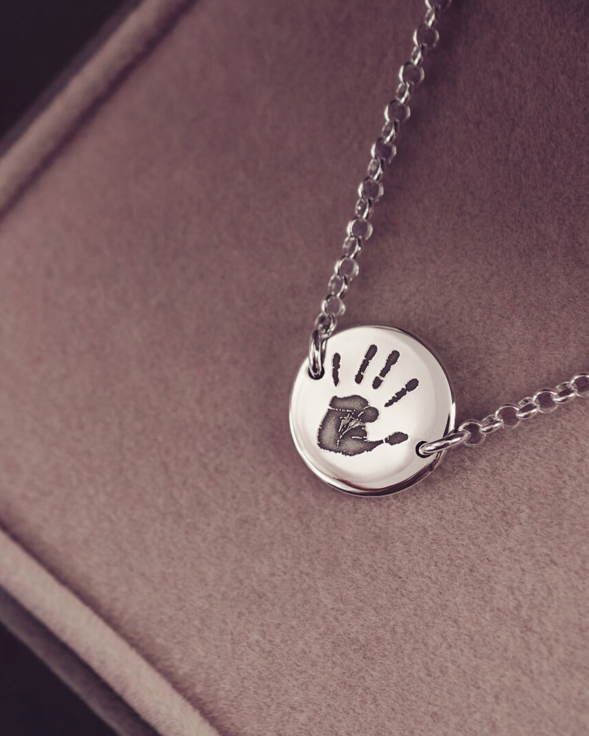 A round silver pendant bracelet featuring a detailed engraved handprint, displayed on a delicate silver chain. The necklace rests in an elegant, soft velvet jewellery box