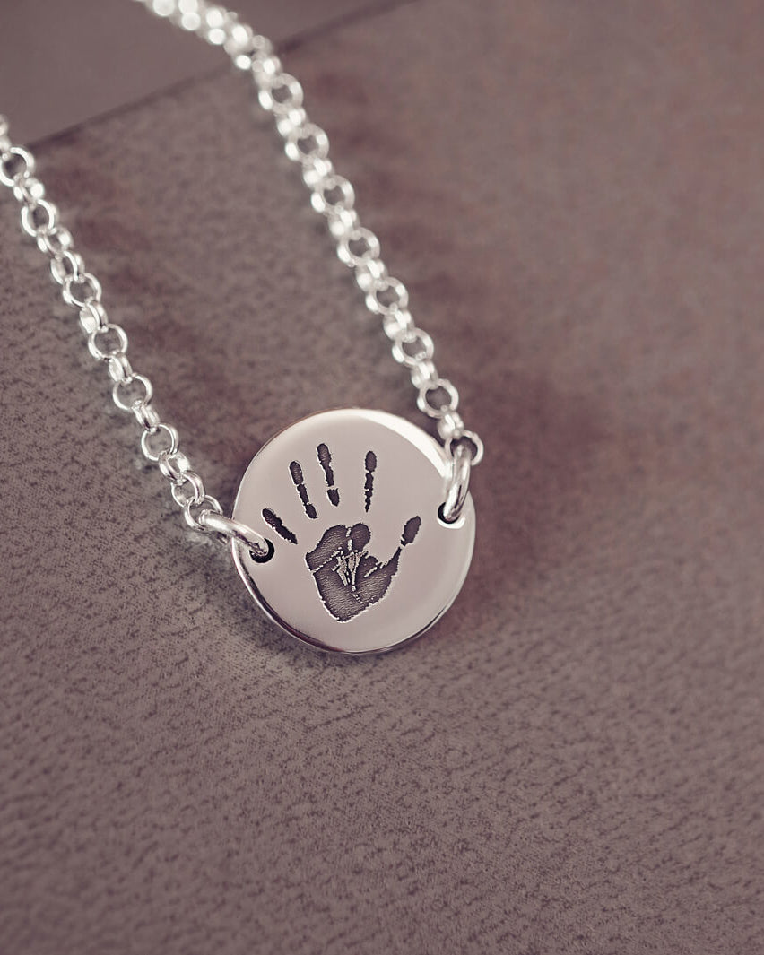 A round silver pendant bracelet featuring a detailed engraved handprint, displayed on a delicate silver chain. The necklace rests in an elegant, soft velvet jewellery box