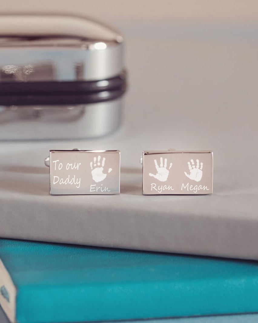 stainless steel rectangular cufflinks with three handprints and names, as well as a short message of 'To Our Daddy' engraved onto the front of the cufflinks. Displayed on a blue and grey background