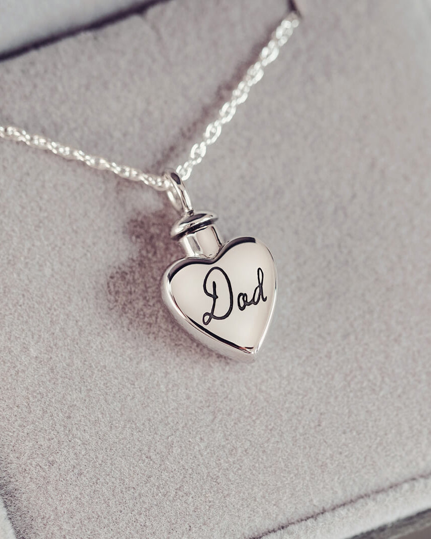 A silver, heart shaped self-fill urn necklace, on a silver chain, with a handwritten message of 'Dad' engraved into the front. Displayed on a grey background