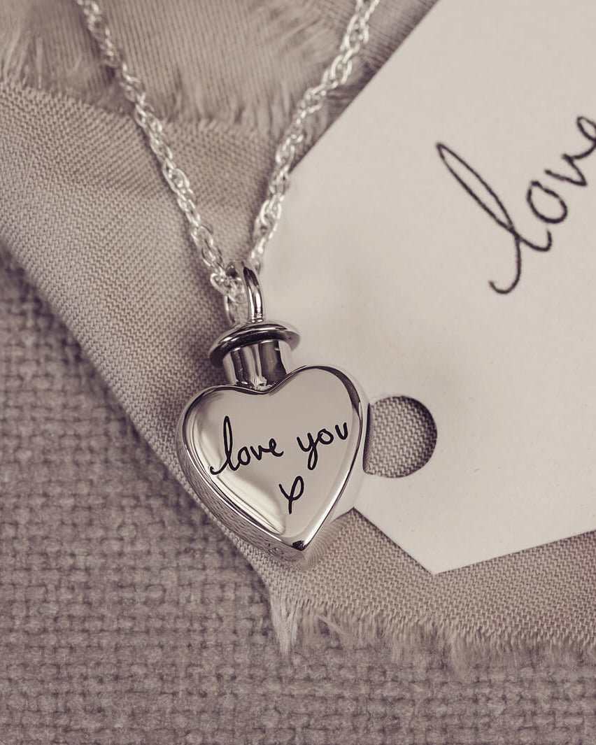A silver, heart shaped self-fill urn necklace, on a silver chain, with a handwritten message of 'love you x' engraved into the front. Displayed on a neutral background